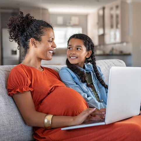 Pregnant black woman working on laptop while sitting on sofa and smiling to daughter. Cheerful expecting mother and girl relaxing on couch and using laptop to book vacation. Happy woman with baby bump and daughter working on computer from home during maternity leave.
