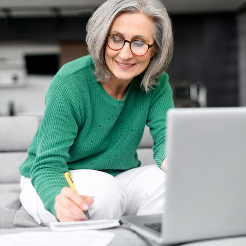 Confident mature woman using laptop computer for remote work, watching webinar and taking notes sitting at home. Contemporary senior female online teacher holding video conference and making marks