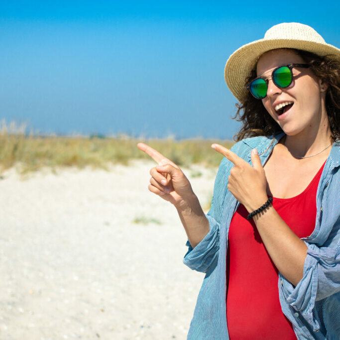 Happy beautiful girl pointing finger at blank copy space for advertising text or promotional, standing on beach, wearing red swimsuit bikini, sunglasses, straw hat. Travel summer sea resort vacation
